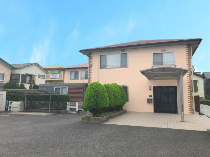 介護施設の建物外観。ベージュと茶色の2階建て、駐車場と植栽完備。